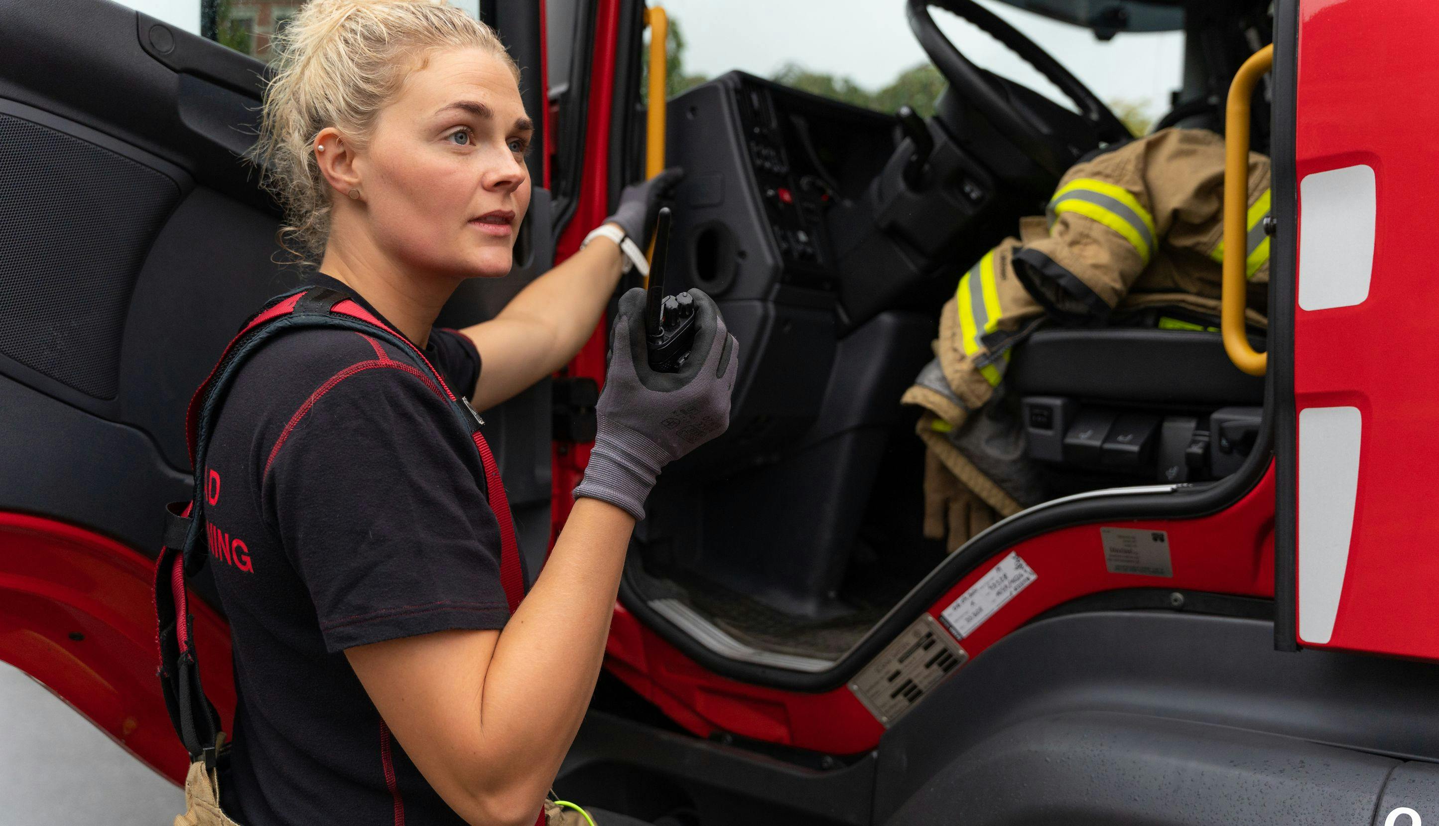 Kvinnelig brannkonstabel står ved brannbil og snakker i walkie talkie. 