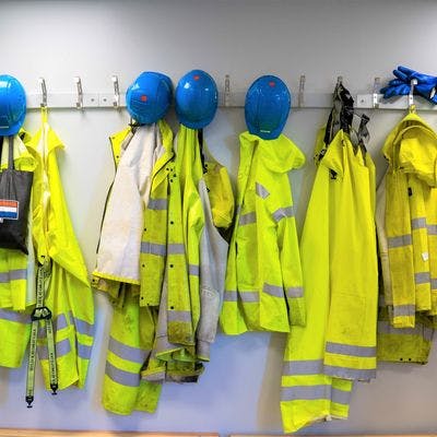 Flere gule arbeidsklær og blå hjelmer henger side om side i en garderobe på en arbeidsplass.