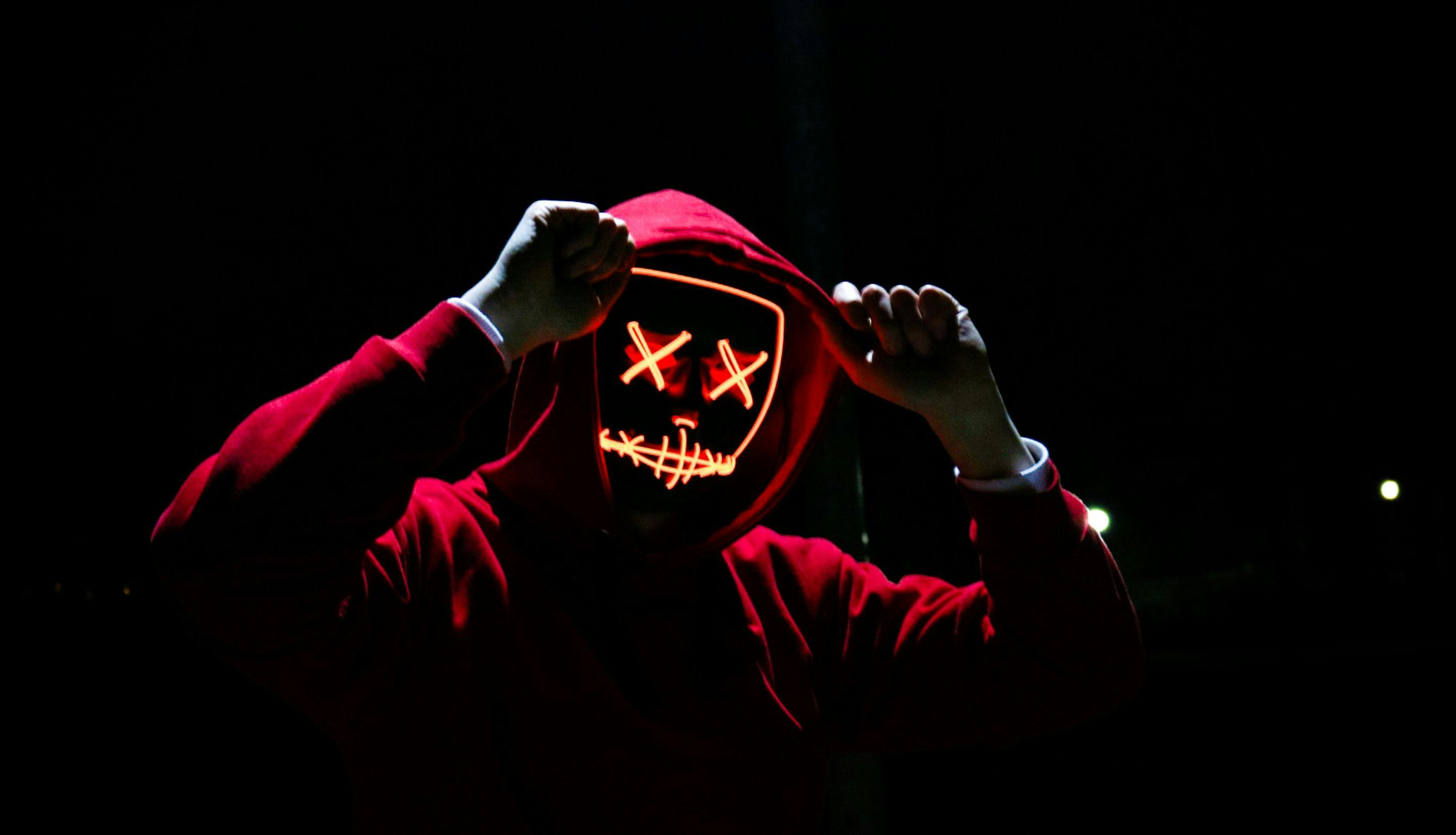 Mannlig skikkelse med rød hettegenser og en selvlysende Halloweenaktig maske ser inn i kameraet.