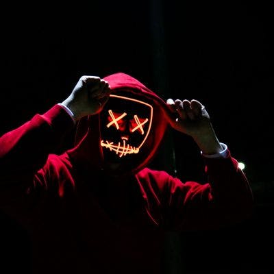 Mannlig skikkelse med rød hettegenser og en selvlysende Halloweenaktig maske ser inn i kameraet.