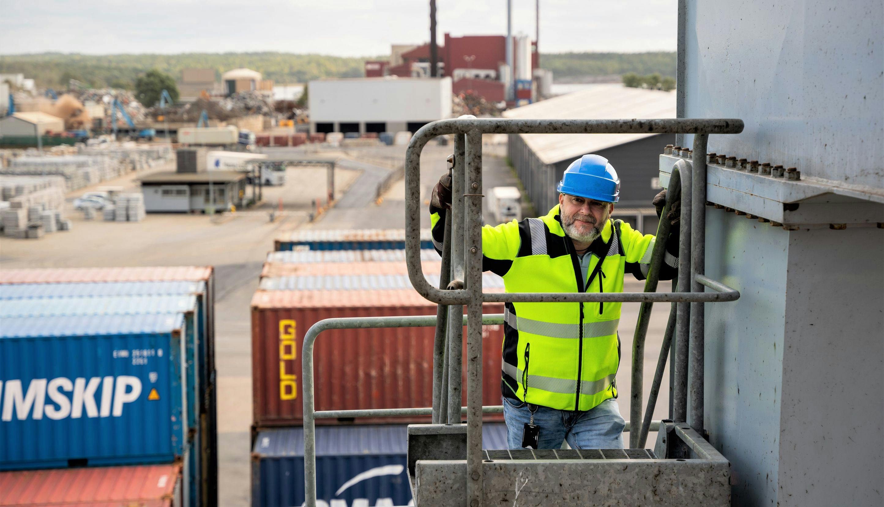 Mann med refleksvest og hjelm klatrer opp en ekstern trapp på et industribygg i en havn. I bakgrunnen synes containere. 