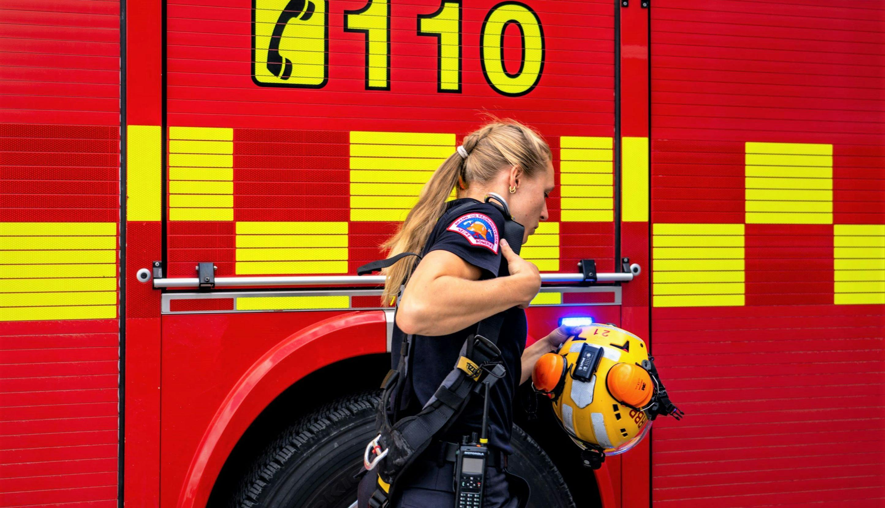 Kvinnelig brannkonstabel holder en hjelm mens hun går forbi en brannbil.