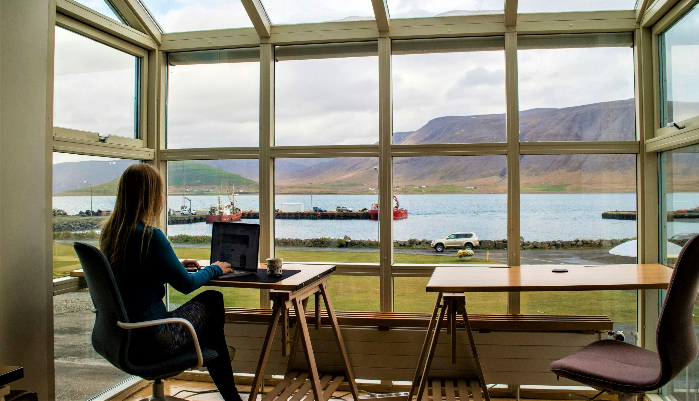 Kvinne som sitter på hjemmekontor ved vindu med utsikt mot fjord. 