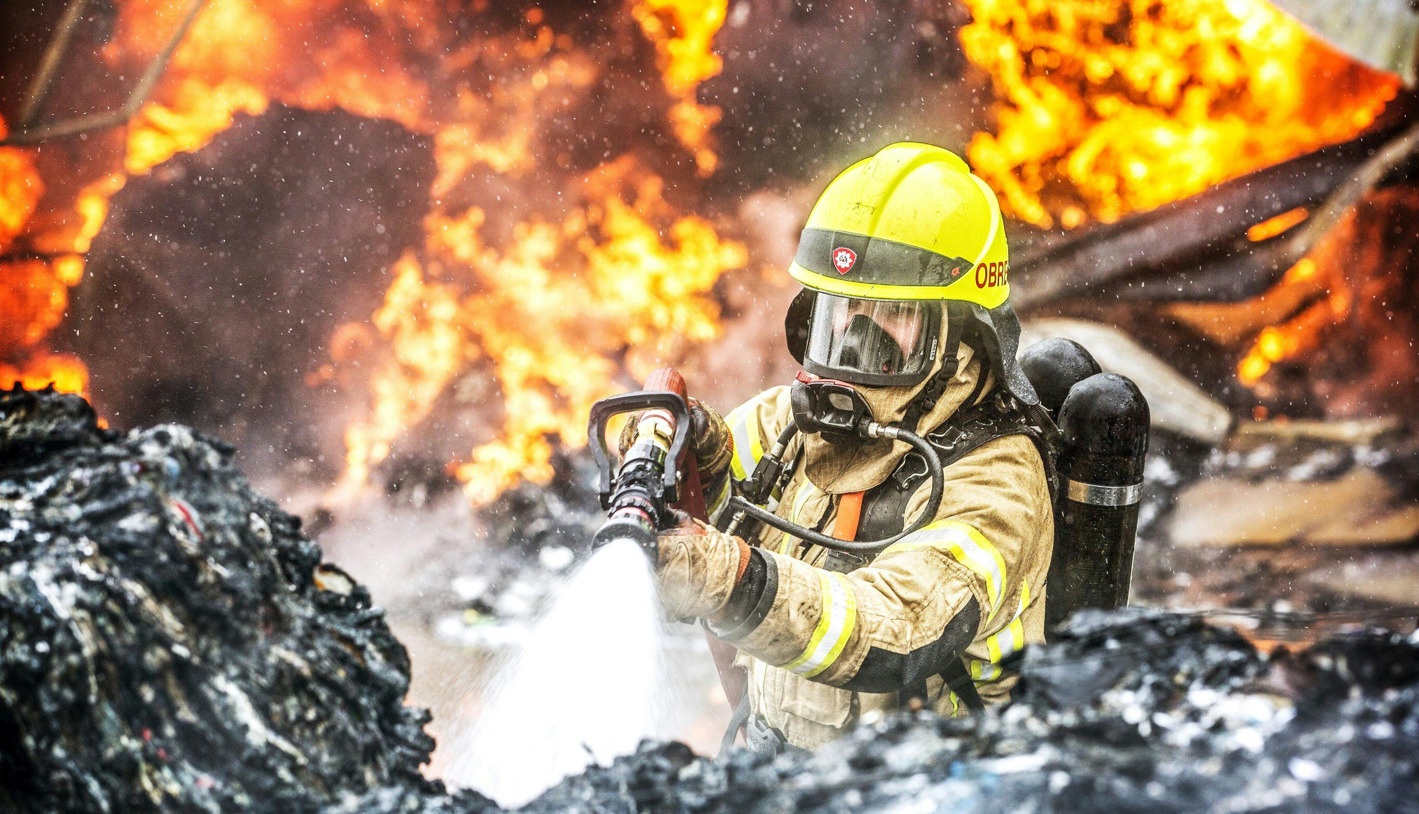 Brannbetjent i fullt utstyr spruter vann på avfall i brann. 