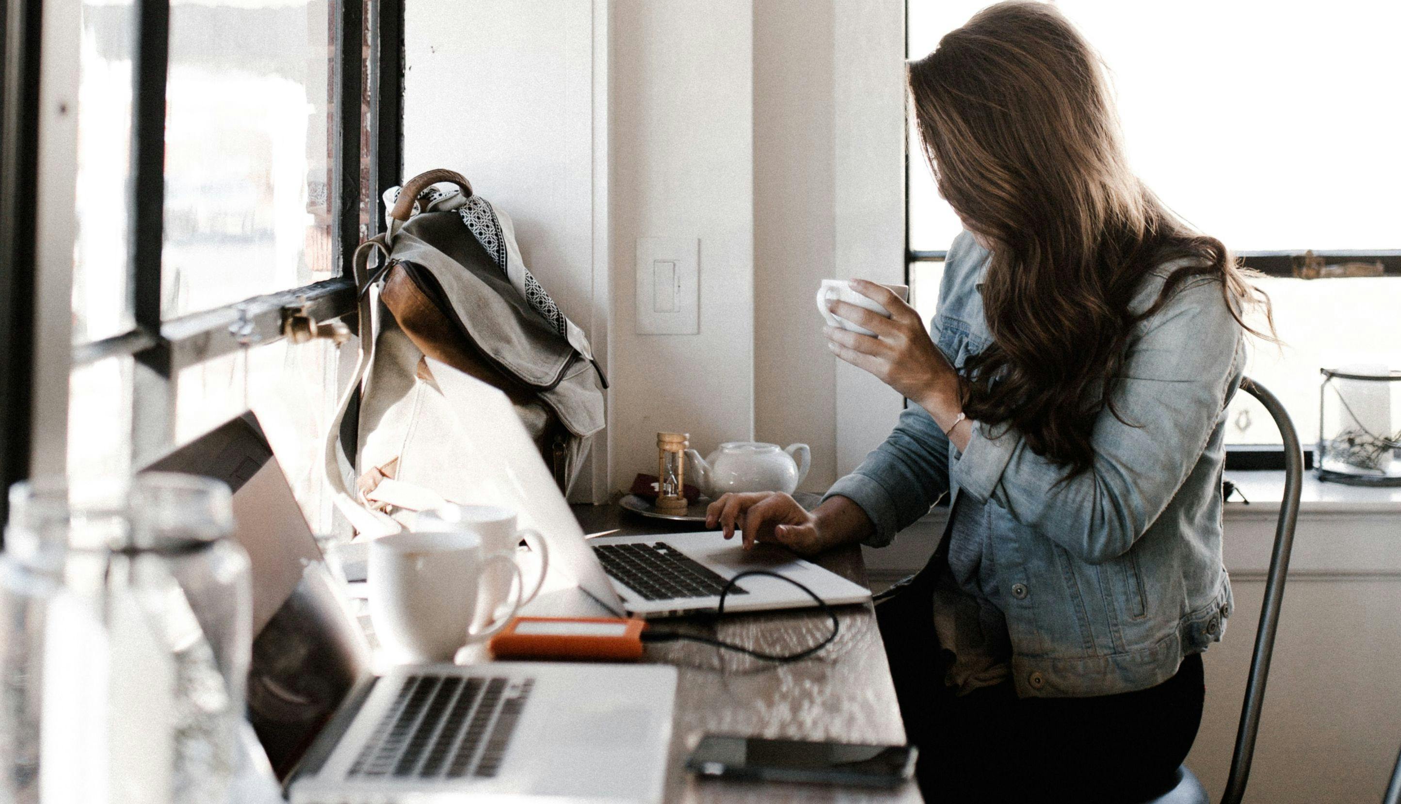 Kvinne som sitter med kopp og jobber ved datamaskin.