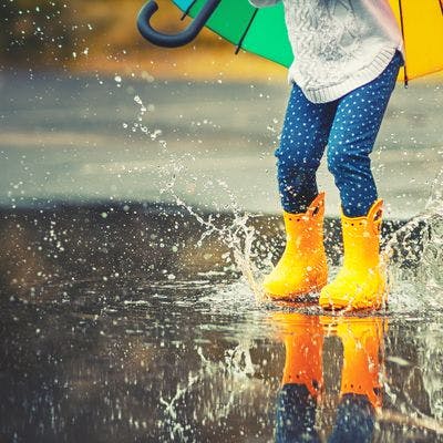 Barn med fargerik paraply og gule støvler hopper i vannpytt så det plasker. 